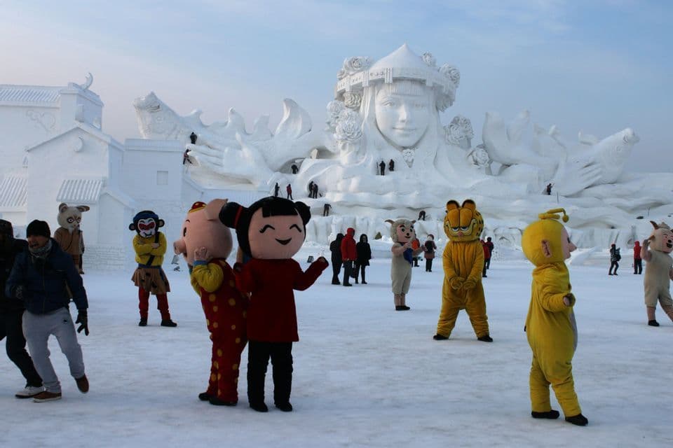 Persone in costumi di personaggi dei cartoni animati si trovano su un campo innevato con sculture di neve imponenti durante un festival invernale.
