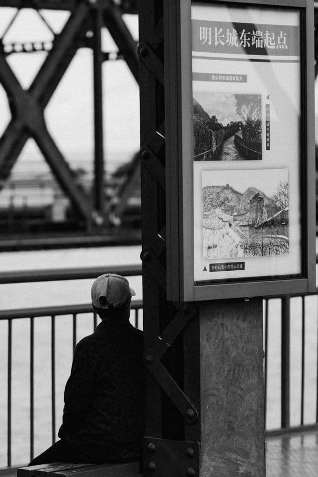 Una persona con un berretto siede su una panchina, vista di spalle, accanto a un cartello informativo con un ponte sullo sfondo. Foto in bianco e nero.