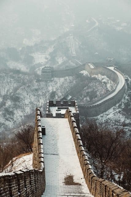 Una vista da una torre di guardia mostra la Grande Muraglia Cinese coperta di neve mentre serpeggia tra le colline nebbiose e alberate.