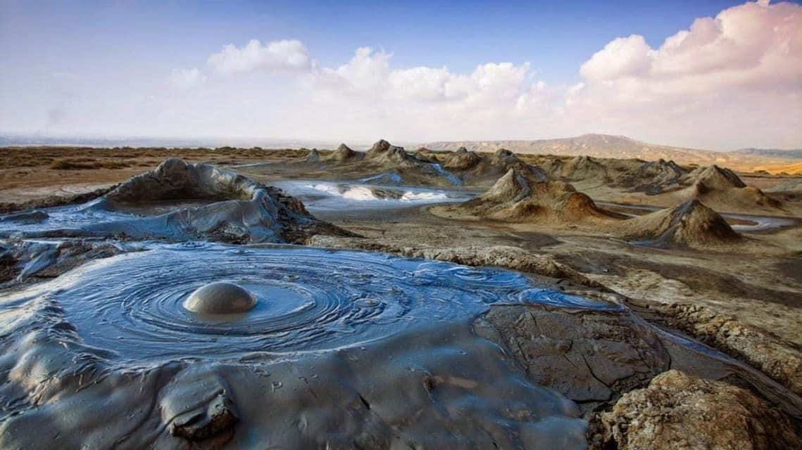 Ein blubbernder Schlammvulkan erzeugt Kräuselungen im Vordergrund einer trockenen Landschaft mit zahlreichen weiteren kleinen Schlammvulkanen.