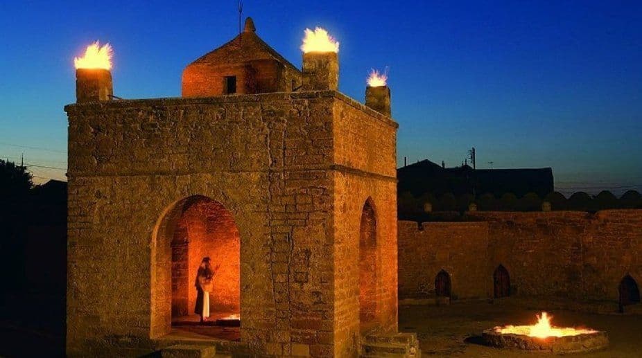 Ein steinerner Feuertempel in der Dämmerung, mit Flammen auf seinem Dach und in einer Feuerstelle im Hof. Eine Person ist in einem beleuchteten Torbogen sichtbar.