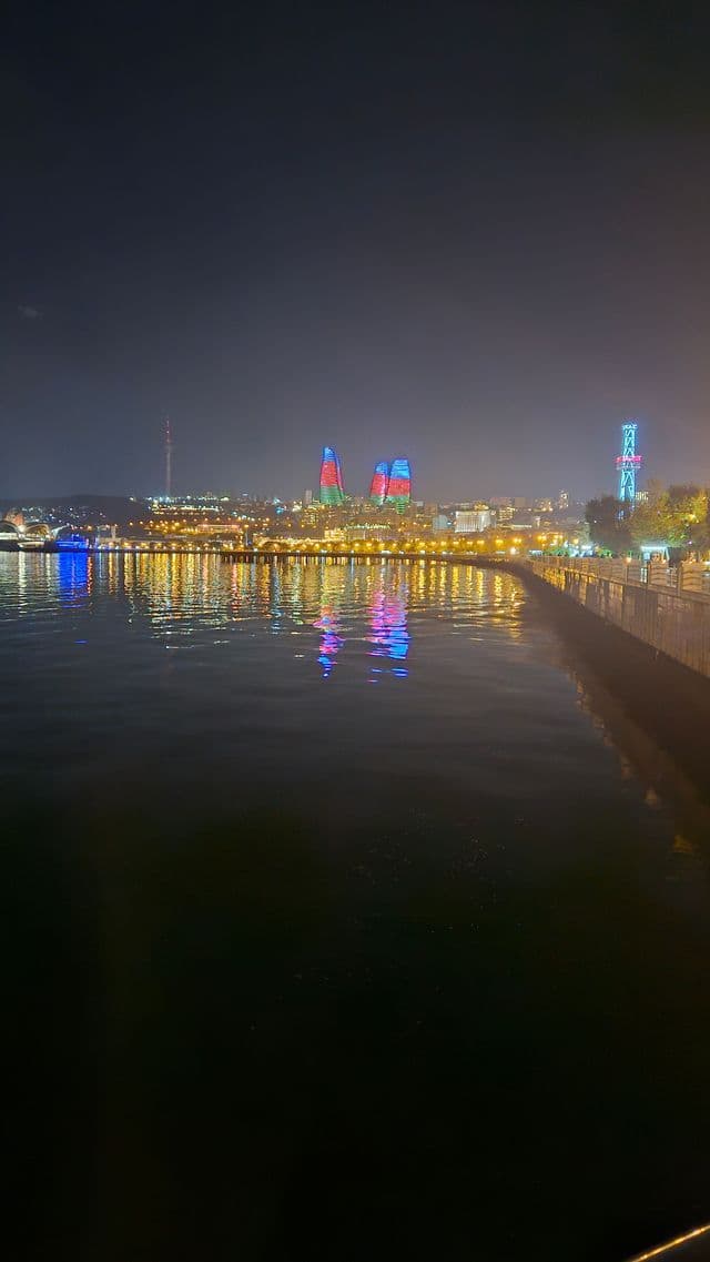 Eine nächtliche Stadtsilhouette, in der zwei flammenförmige Türme mit roten, blauen und grünen Lichtern sich auf dem stillen Wasser spiegeln.