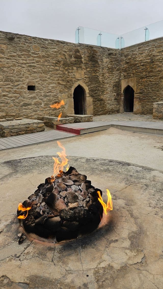 Ein Feuer brennt in einer zentralen Steingrube in einem alten Innenhof, der von Steinmauern mit gewölbten Torbögen umschlossen ist.