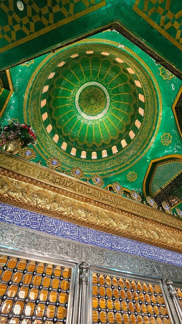 Eine grüne Mosaikkuppel mit goldener Kalligrafie, aus der Untersicht fotografiert, über prächtigen goldenen, blauen und silbernen Friesen und einem Gitter.