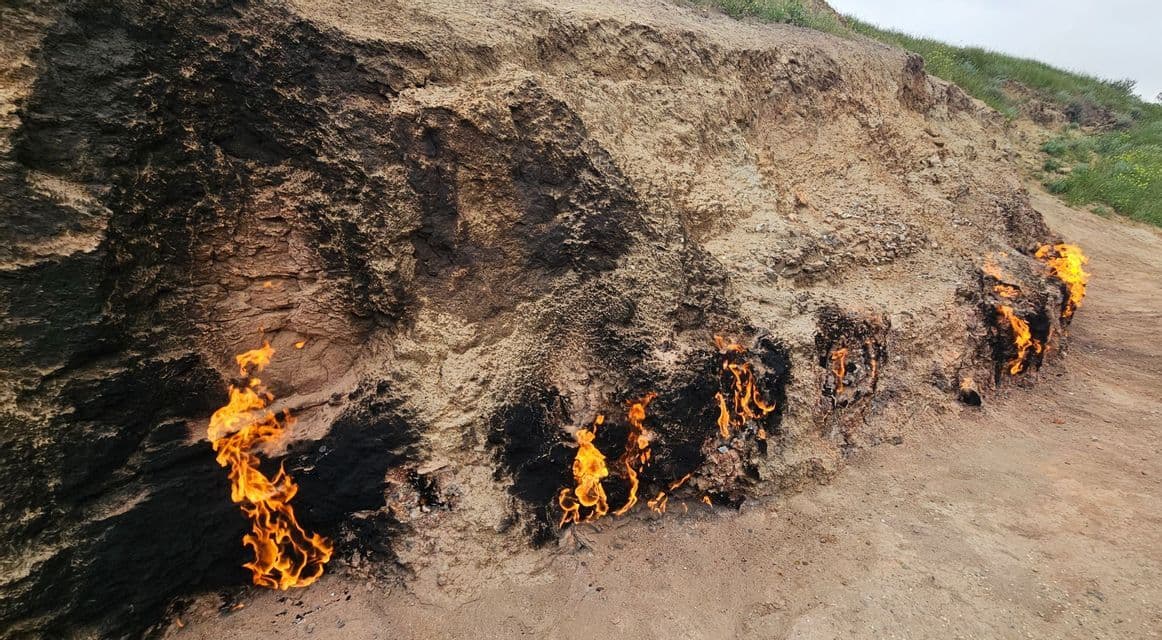 Flammen brennen an mehreren Stellen am Fuße eines versengten, felsigen Hügels unter bedecktem Himmel.