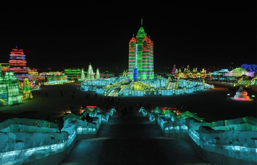 Grandi strutture ed edifici di ghiaccio illuminati da luci colorate di notte, con persone che camminano sulla neve sottostante.