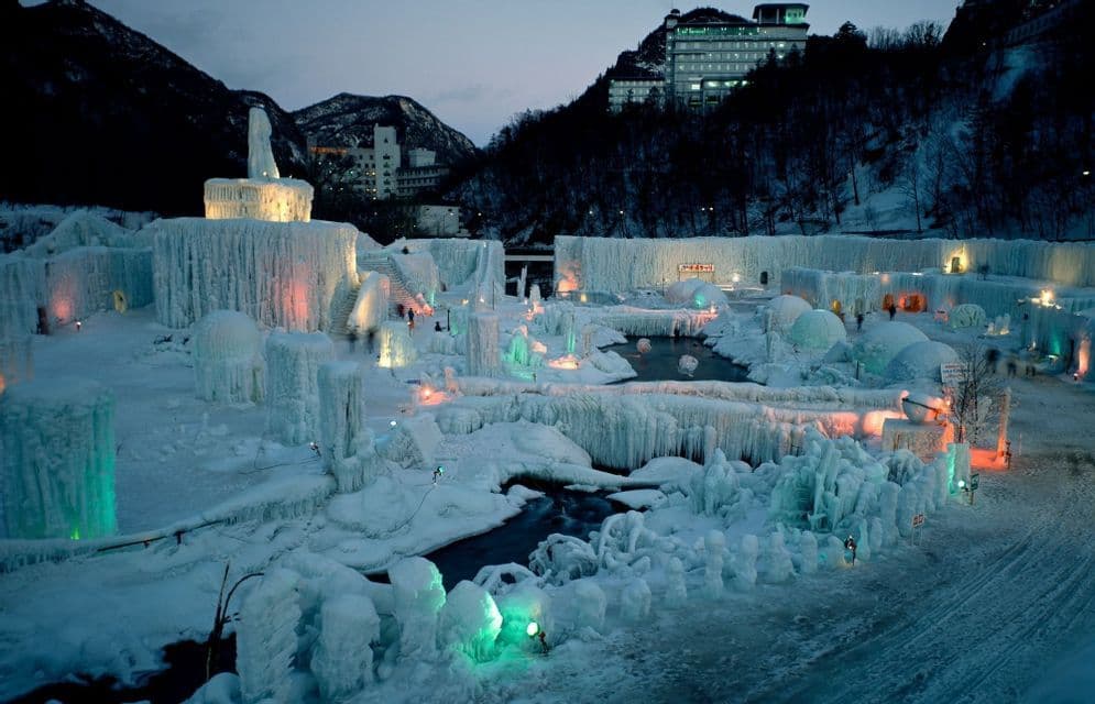 Un grande festival del ghiaccio con strutture e sculture ghiacciate illuminate da luci colorate al tramonto in un ambiente montano innevato.