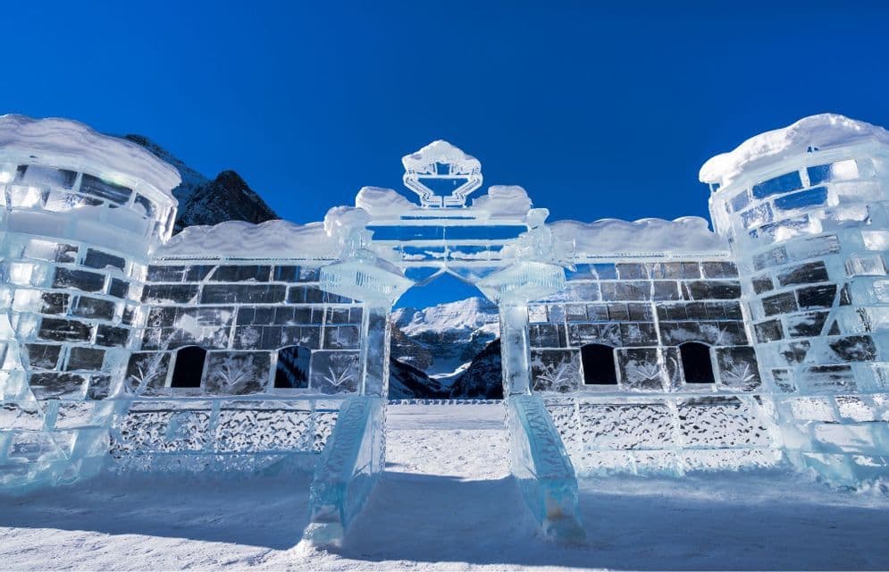 Un grande castello di ghiaccio con un portale ad arco si erge nella neve, incorniciando montagne lontane sotto un cielo azzurro e limpido.