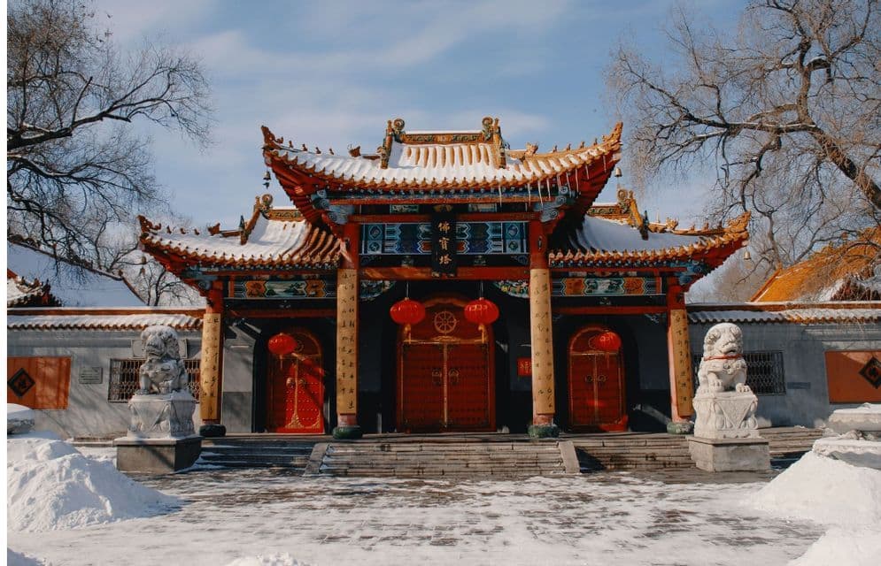 Un ingresso di tempio est-asiatico ornato con lanterne rosse e un tetto coperto di neve si trova in un cortile innevato.