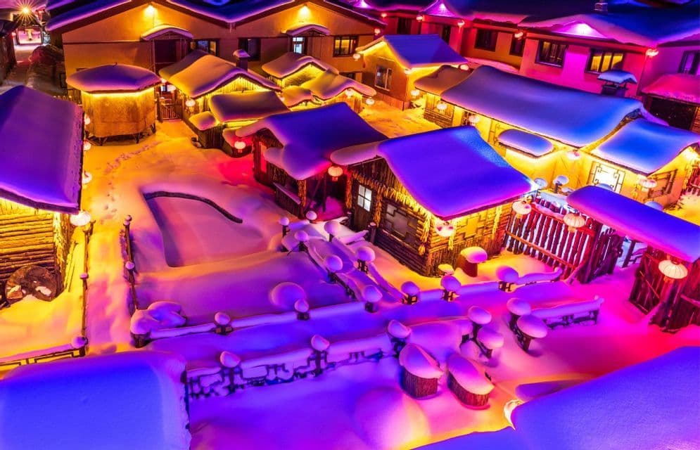Veduta aerea di un villaggio innevato di notte, con edifici illuminati da luci colorate gialle e viola.