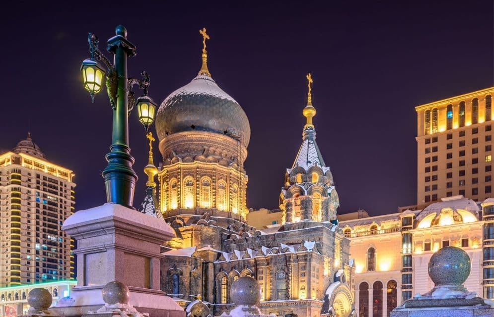 Una cattedrale ornata, con cupole a cipolla, coperta di neve, è illuminata di notte accanto a un lampione acceso e a edifici moderni della città.