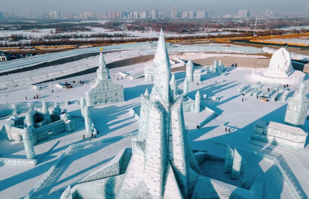Maestose sculture di ghiaccio di castelli ed edifici riempiono un paesaggio innevato sotto un cielo limpido, con lo skyline di una città in lontananza.