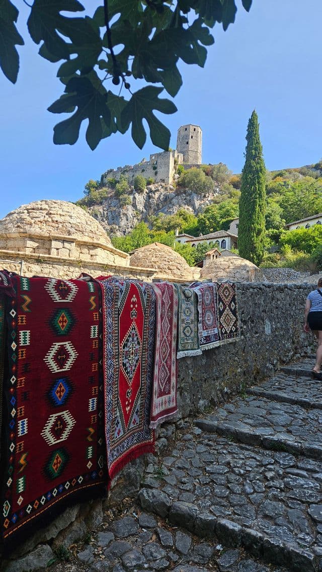 Bunte gemusterte Teppiche hängen an einer Steinmauer entlang eines Kopfsteinpflasterwegs, mit einer Steinfestung auf einem grünen Hügel im Hintergrund.