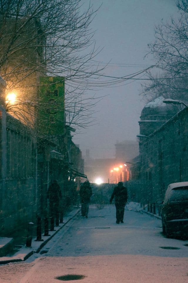 Trois personnes marchent dans une rue de ville enneigée au crépuscule, sous une forte chute de neige.