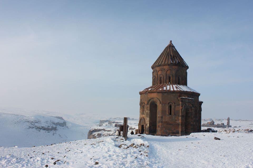 Une église historique en pierre polygonale au toit conique se dresse sur une colline enneigée au milieu de ruines antiques.