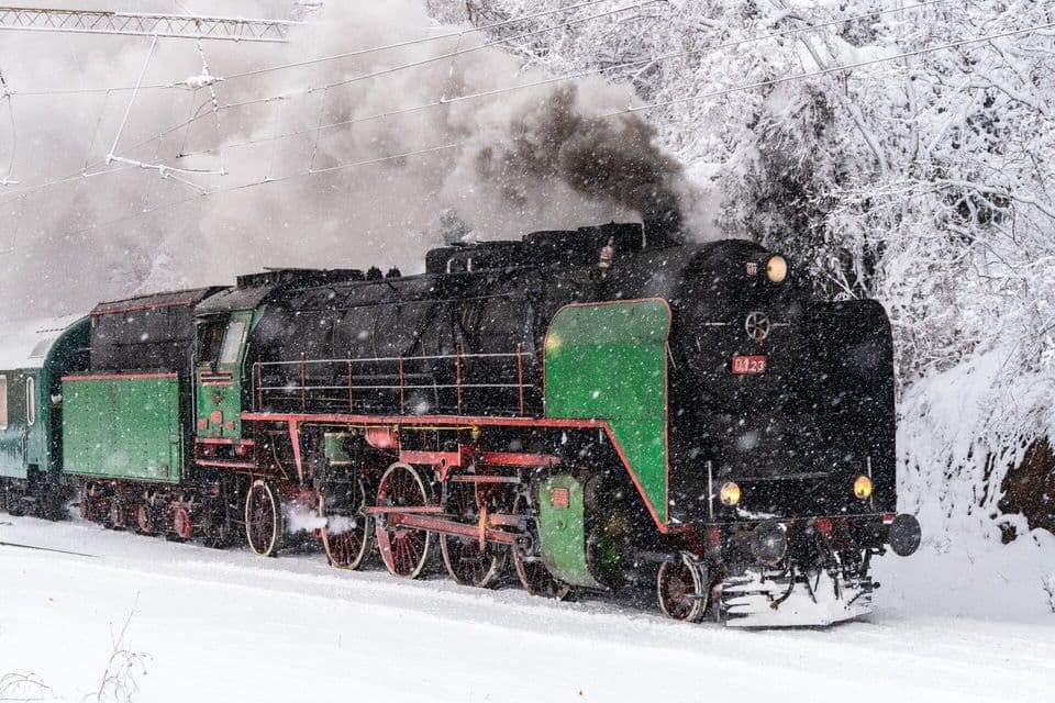 Une locomotive à vapeur vintage, noire et verte, se déplace sur les rails sous une forte chute de neige, émettant une fumée sombre.