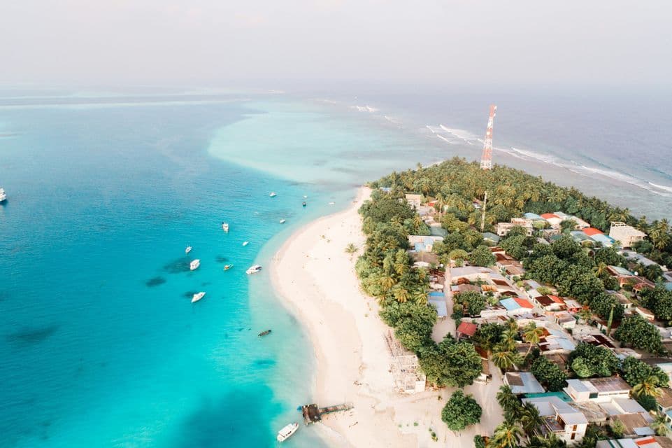 An aerial view of a tropical island peninsula with a village, a white sand beach, and boats in the turquoise water.
