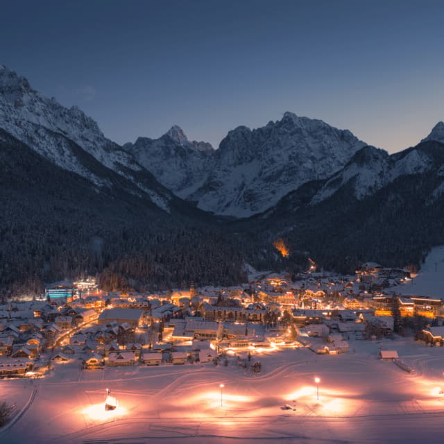 Un villaggio innevato che brilla di luci in una valle di montagna al tramonto sotto un cielo sereno.