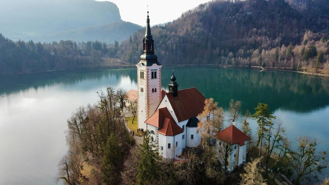 Veduta aerea di una chiesa bianca con un campanile su una piccola isola in un lago tranquillo, circondata da colline boscose.
