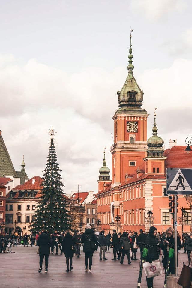 Une foule se promène sur une place historique européenne, ornée d'un grand sapin de Noël et d'un imposant bâtiment à tour horloge rouge.
