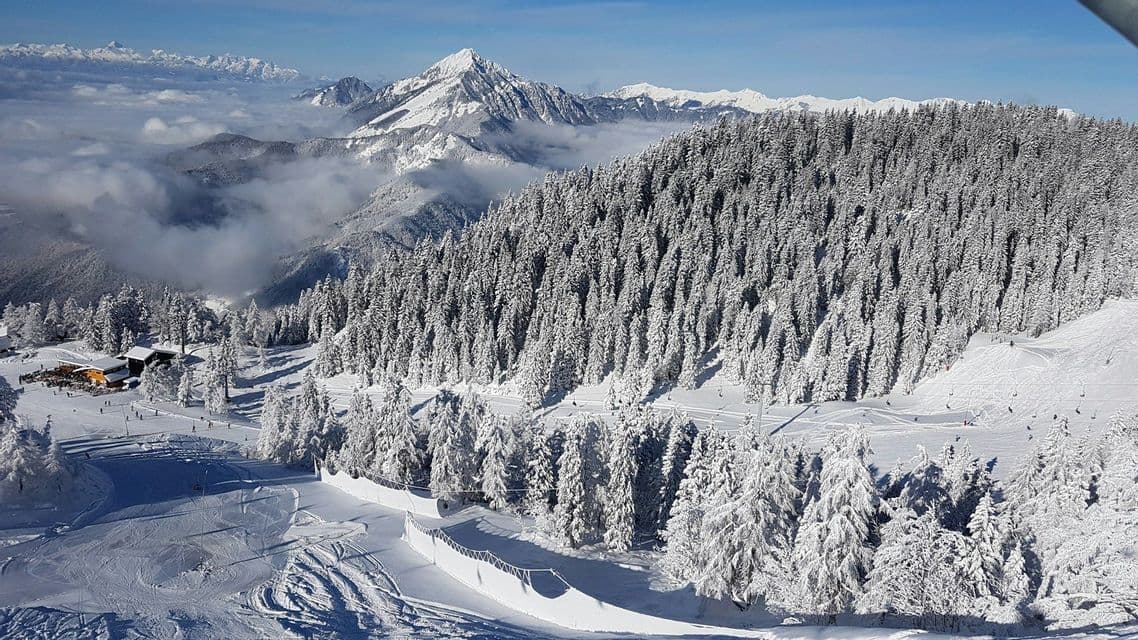 Una vista panoramica su una stazione sciistica con pinete innevate, piste da sci attive e una catena montuosa che si innalza sopra le nuvole.