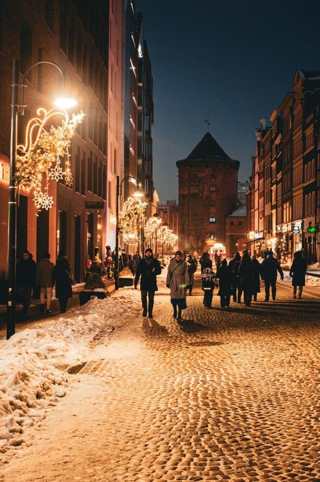 Des gens marchent sur une rue pavée la nuit, avec de la neige sur les côtés et des lumières festives décorant les lampadaires.