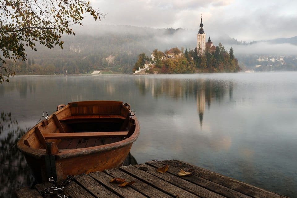 Una barca di legno è ormeggiata a un molo su un lago nebbioso, di fronte a un'isola lontana con una chiesa il cui campanile si riflette nell'acqua.