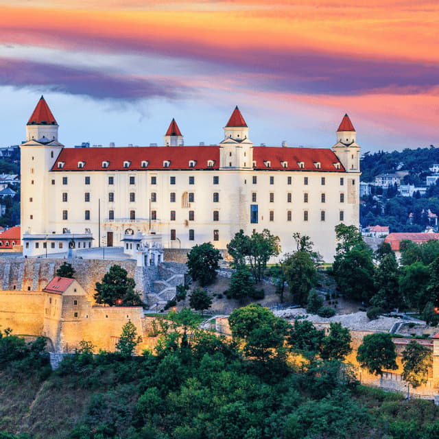 Un grande castello bianco illuminato con tetti rossi si erge su una collina con fortificazioni e alberi, durante un tramonto colorato.