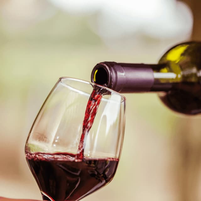Vino rosso versato da una bottiglia scura in un calice trasparente, su uno sfondo sfocato.