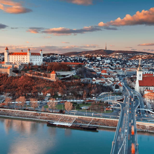 Un castello bianco su una collina domina una città europea con un fiume e un ponte trafficato al tramonto.