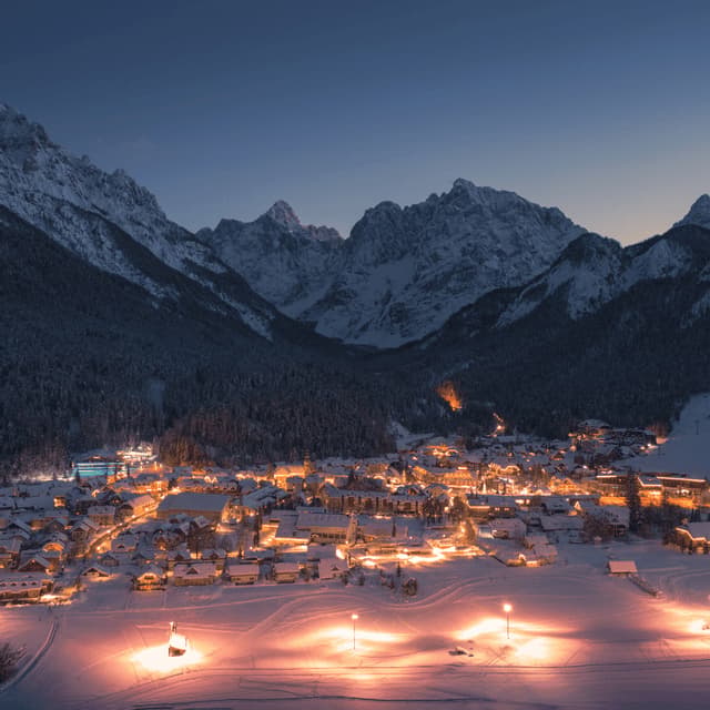 Un villaggio innevato brilla di luci calde in una valle di montagna al crepuscolo, sotto imponenti cime innevate.
