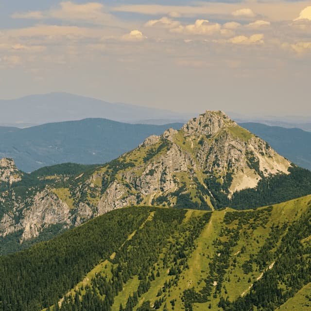 Una cima rocciosa si erge sopra verdi colline boscose sotto un cielo parzialmente nuvoloso.