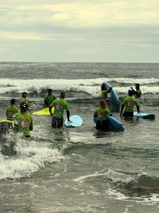 Un gruppo WeRoad in magliette verdi abbinate si prepara per una lezione di surf, in piedi con le tavole nell'oceano mosso.