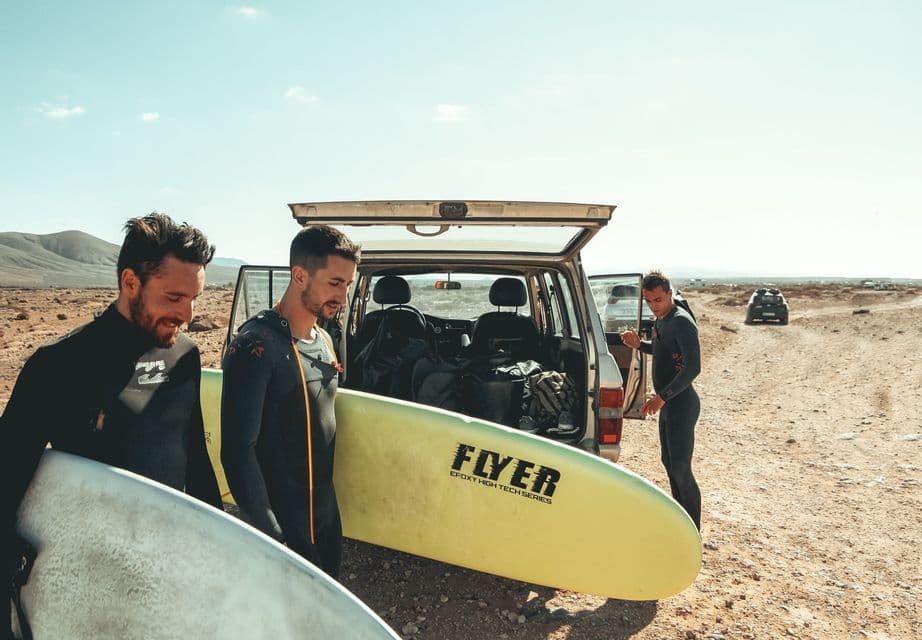 Un viaggio di gruppo WeRoad di tre uomini in muta che prendono le loro tavole da surf dal retro di un SUV in un paesaggio secco e soleggiato.