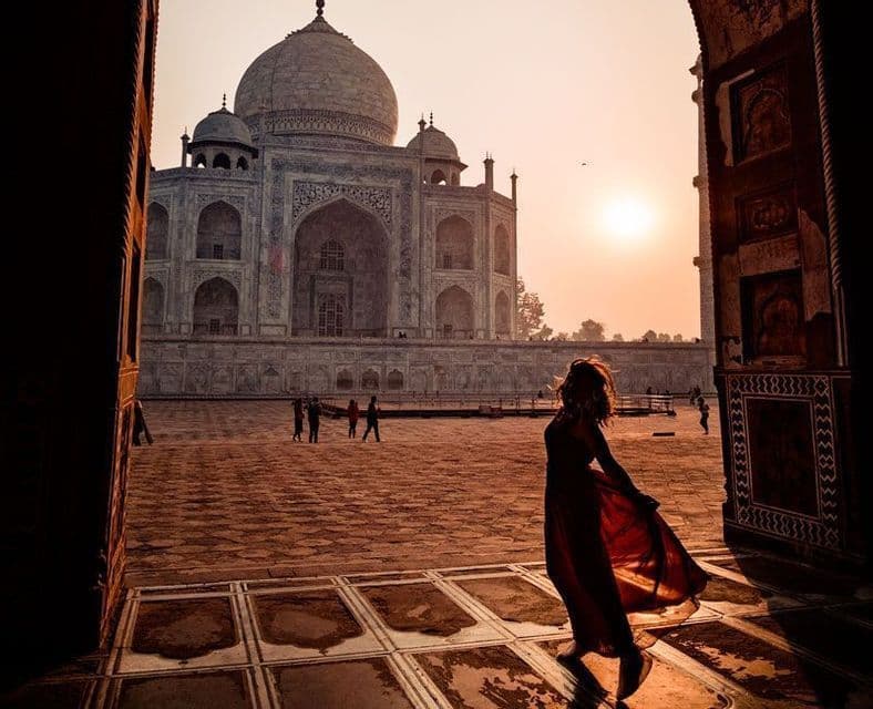 Una donna in abito rosso si staglia in silhouette in un arco di pietra, ammirando il Taj Mahal all'alba.