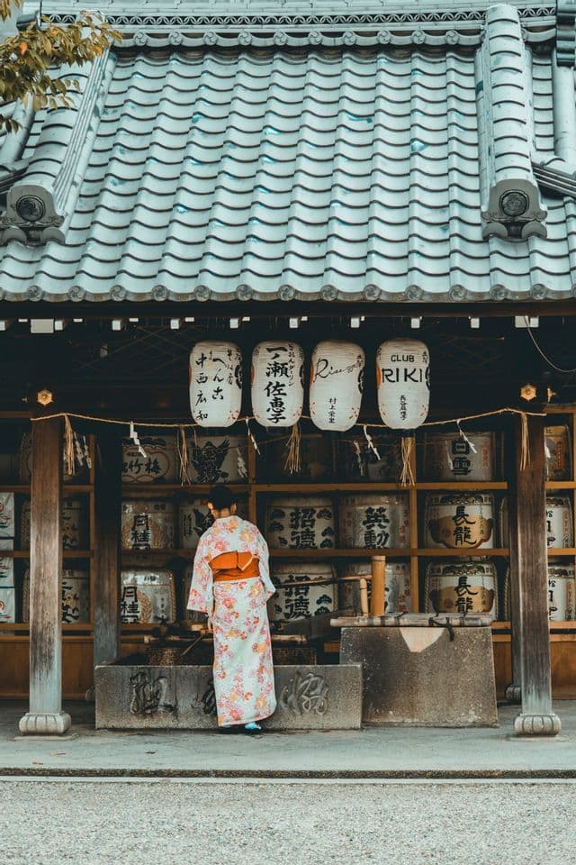 Une personne vue de dos, portant un kimono fleuri, se tient devant un temple japonais traditionnel orné de lanternes suspendues.