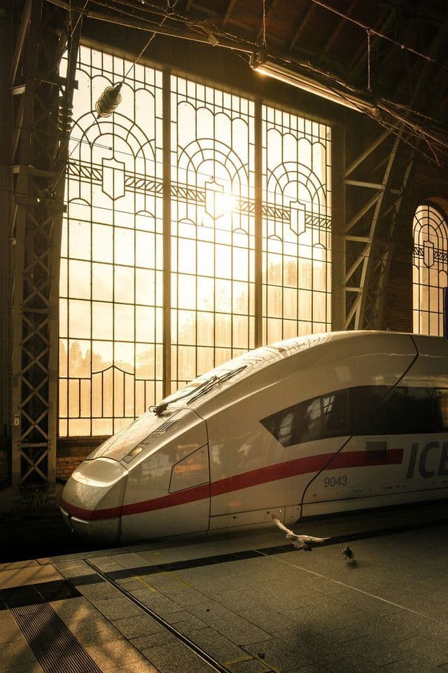 Un treno bianco ad alta velocità fermo in una stazione, con la luce intensa del sole che filtra attraverso grandi finestre ad arco sullo sfondo.