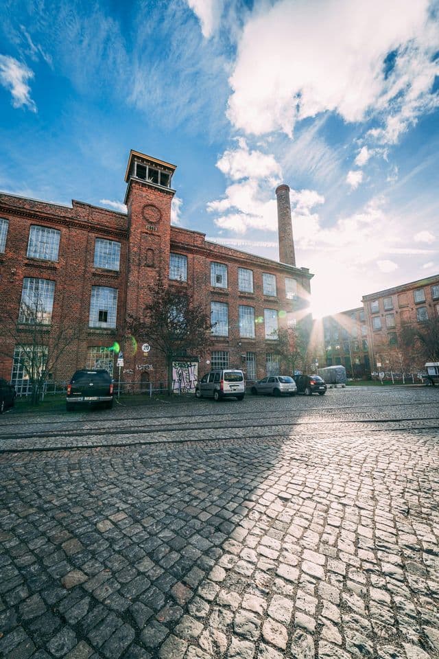 Una vista dal basso di un edificio industriale in mattoni rossi con una ciminiera, ripreso da un cortile acciottolato sotto un cielo azzurro brillante.