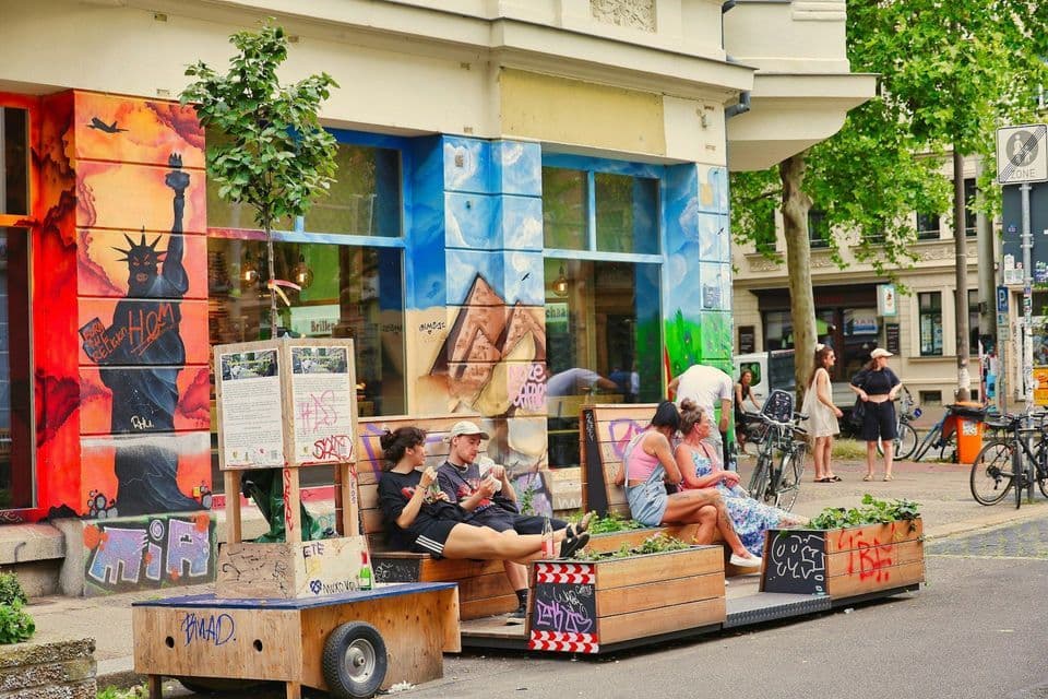 Un gruppo di persone è seduto su un parklet in legno in una strada di città, davanti a un edificio ricoperto di murales colorati.