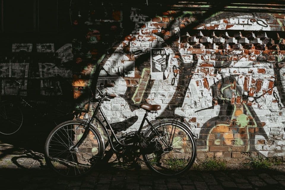 Una bicicletta nera è parcheggiata su una strada acciottolata, proiettando un'ombra su un muro di mattoni rovinato e coperto di graffiti.