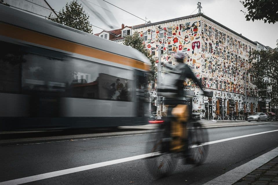 Un ciclista sfocato dal movimento sfreccia accanto a un tram su una strada di città, con un edificio adornato da murales colorati sullo sfondo.