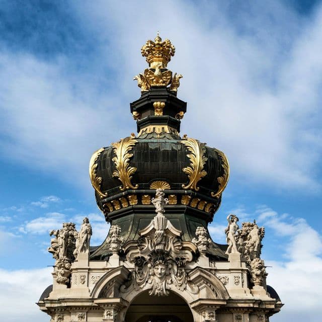 Una grande cupola a forma di corona, nera e dorata, riccamente ornata, sorge su un arco in pietra con sculture, contro un cielo azzurro.