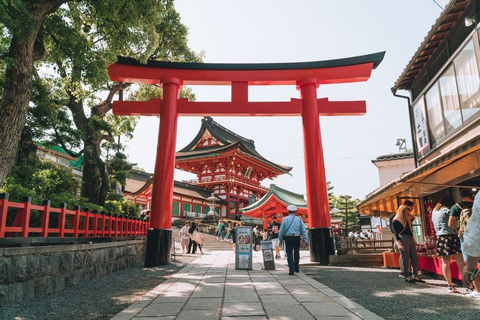 Ein großes rotes Torii-Tor umrahmt einen traditionellen japanischen Tempel, während Menschen an einem sonnigen Tag einen Steinpfad entlanggehen.