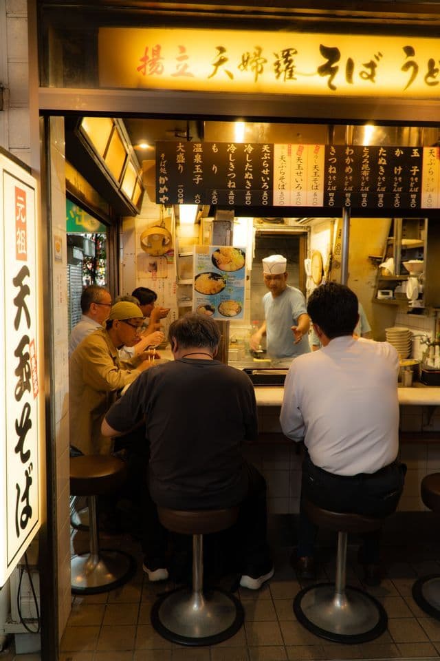 Männer essen Nudeln an der Theke eines hell beleuchteten japanischen Restaurants, während ein Koch in der Küche zu sehen ist.