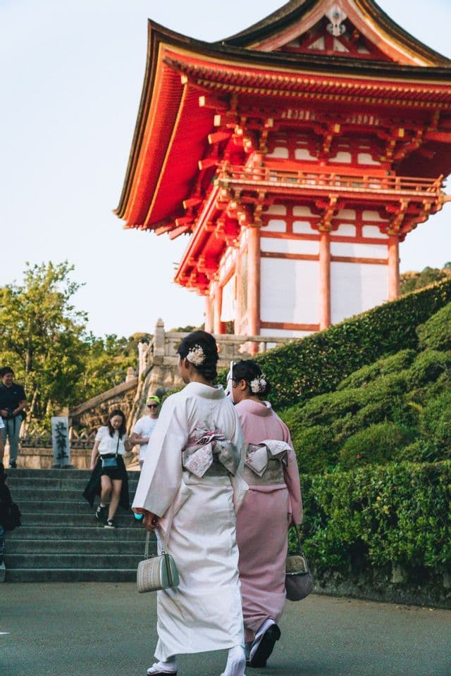 Zwei Frauen in traditionellen Kimonos gehen auf einem Pfad zu einer roten japanischen Pagode auf einem Hügel, von hinten gesehen.