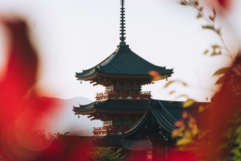 Eine mehrstöckige japanische Pagode mit einem dunklen Ziegeldach ist durch verschwommene rote Blätter vor einer dunstigen Bergkulisse zu sehen.
