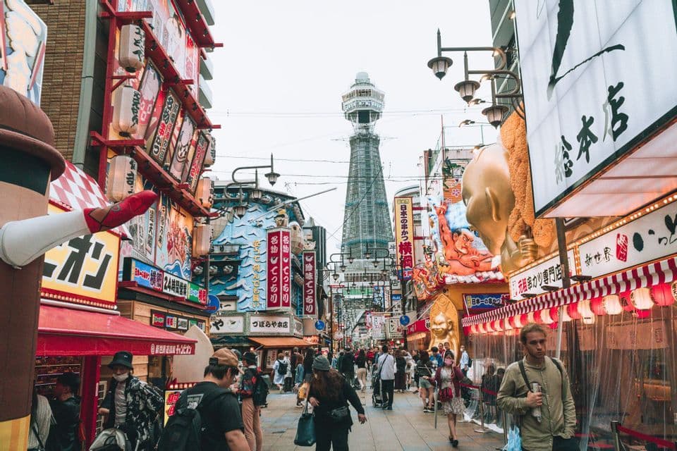 Eine belebte Straße in einer japanischen Stadt mit vielen bunten Schildern und Laternen, mit einem hohen Aussichtsturm im Hintergrund.
