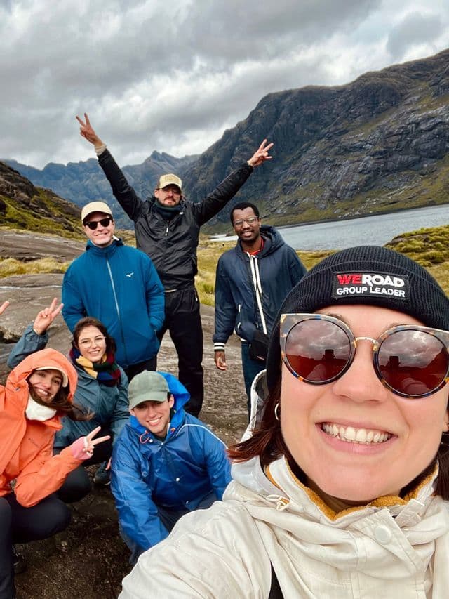 Eine WeRoad-Gruppenreise posiert für ein Selfie in einer bergigen Landschaft an einem See unter einem bewölkten Himmel.