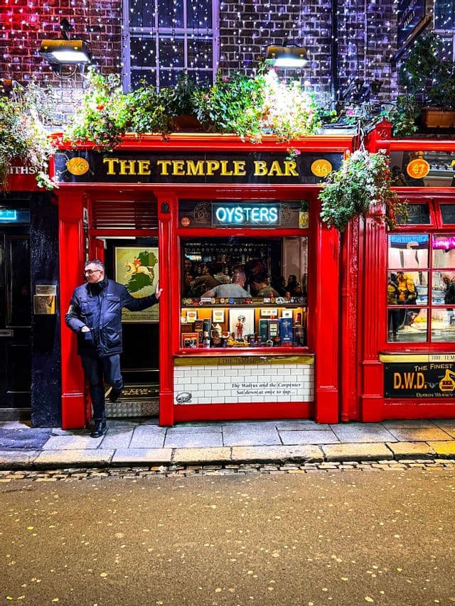 Ein Mann in dunkler Jacke posiert im Eingangsbereich der The Temple Bar, die nachts eine hell erleuchtete rote Fassade hat.