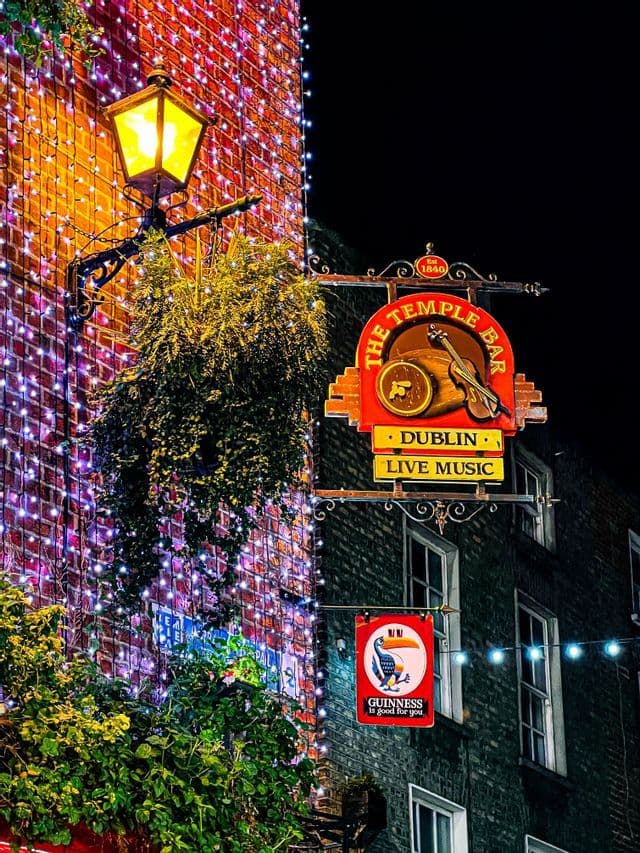 Das beleuchtete Schild der Temple Bar in Dublin hängt nachts an einer Backsteinfassade mit Lichterketten und einer Straßenlaterne.